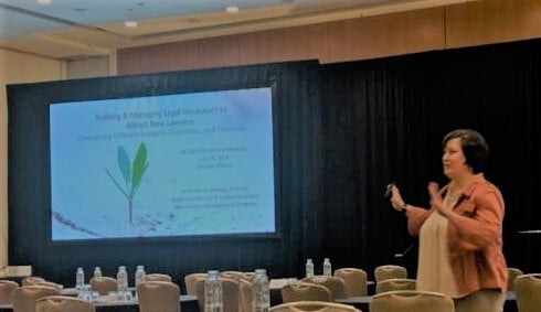 Anne-Marie delivering Building & Managing Legal Incubators to Attract New Lawyers: Considering Different Budgets, Objectives, and Timeline as a keynote at ACLEA's 55th Annual Meeting in 2019. The program's title slide is projected on the screen behind her.